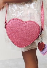 Pink heart-shaped crossbody bag made of glittery material, featuring a silver chain with three smaller heart-shaped charms in different colors.