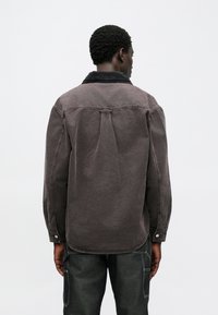 Šedá džínová košile s černým manžestrovým límcem, s knoflíkovými detaily a pohodlným střihem. Má záševky na zádech pro větší volnost pohybu.