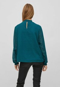 Blusa teal, texturizzata, con maniche lunghe e trasparenti, un sottile motivo a zigzag, chiusura a chiave sul retro e polsini elasticizzati. Abbinata a pantaloni neri.