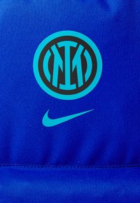Saco de desporto em tecido azul com um logótipo circular preto e turquesa, apresentando letras estilizadas, juntamente com um design do swoosh da Nike em turquesa.