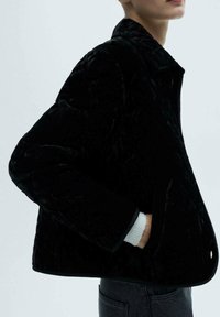 Sort quiltet jakke med høj krave, struktureret velvet-materiale, stiklommer og kontrasterende hvid ærmelinning. Slim fit design.
