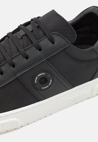 Black sneakers with a mesh and leather upper, rounded toe, textured surface, flat white rubber sole, and a circular logo detail on the side.