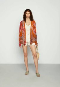 Kimono laranja fluido com padrão paisley, combinado com uma t-shirt creme e calções. Acessorizado com uma carteira de palha e sandálias verdes.
