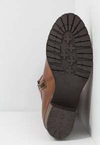 Botte en cuir marron avec semelle en caoutchouc noire montrant des crampons profonds et des motifs en forme de croix, appuyée contre un mur blanc.
