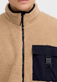 Chaqueta de forro polar beige con cuello alto, cremallera negra y un bolsillo oscuro con detalle de hebilla. Textura suave y diseño informal.