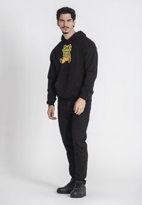 Felpa nera con un grande design grafico giallo-arancione che presenta un personaggio dei cartoni animati. Abbinata a jeans neri e sneakers nere.