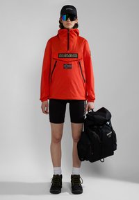 Rode lichtgewicht jas met een voorzak, zwarte accenten, rits en vlagdetail. Zwarte shorts, sportschoenen en een zwarte rugzak met een oprolbare bovenkant.