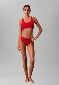 Frau steht barfuß in einem roten Sport-BH von Calvin Klein und passender Unterwäsche vor einem schlichten grauen Hintergrund.
