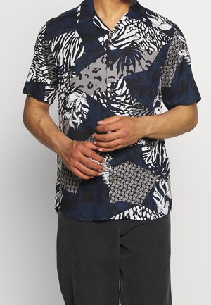 Mand iført en kortærmet skjorte med abstrakte marineblå, hvide og grå tropiske og geometriske mønstre, stående med sammenlagte hænder.