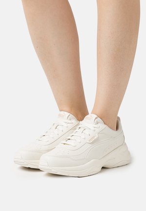 Persoon draagt witte Puma sneakers met dikke zolen op een effen lichte achtergrond, met vooraanzicht en zijaanzicht van de schoenen.