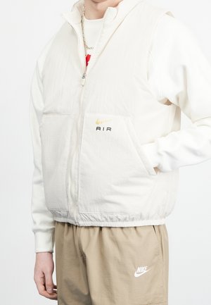 Person iført en hvid dunvest med lynlås med "Nike Air" logo og beige Nike joggingbukser, hånd i vestlommen, kædehalskæde synlig.