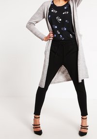 Donna che indossa un top floreale nero, pantaloni neri, un cardigan lungo grigio e tacchi a strappo neri, in posa con una mano sui fianchi.