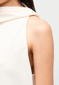 Αμάνικο, απαλό μπεζ τοπ με ντραπαρισμένη λαιμόκοψη. Διαθέτει λεία ύφανση και χαλαρή εφαρμογή, αναδεικνύοντας την περιοχή των ώμων και των βραχιόνων.