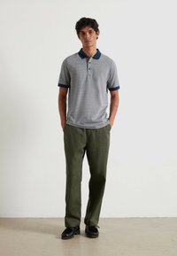 Polo grigio con colletto e polsini blu navy, caratterizzato da un motivo sottile. Abbinato a pantaloni verde oliva e scarpe nere con suola texturizzata.