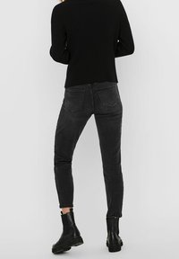 Jean skinny noir avec poches arrière, taille haute et texture lisse, associé à des bottines noires. La tenue présente un design simple et moderne.