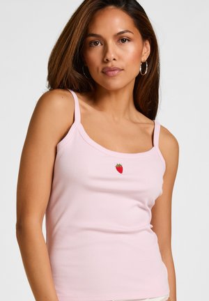 Femme aux longs cheveux bruns portant des boucles d'oreilles en créoles et un débardeur rose avec un petit emblème rouge en forme de fraise sur la poitrine.