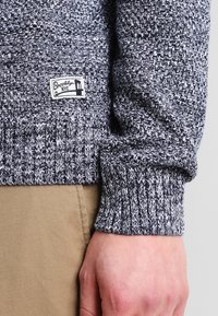 Granatowo-biały dzianinowy sweter z prążkowanymi mankietami, z metką "Brooklyn NYC". Miękka tekstura i widoczny wzór tkany.