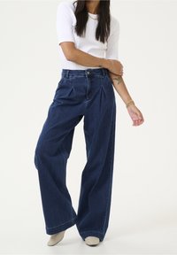 Tummansiniset leveälahkeiset farkut, joissa on pleateja, korkea vyötärö, vyölenkit ja etutaskut, yhdistettyinä tiukkaan valkoiseen toppiin.