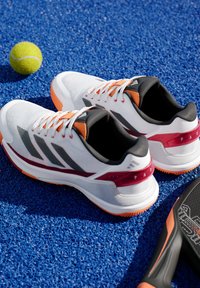 Witte sportschoenen met zwart en grijs gestreepte details, oranje accenten en gestructureerde zolen, geplaatst op een blauwe synthetische ondergrond met een tennisbal.