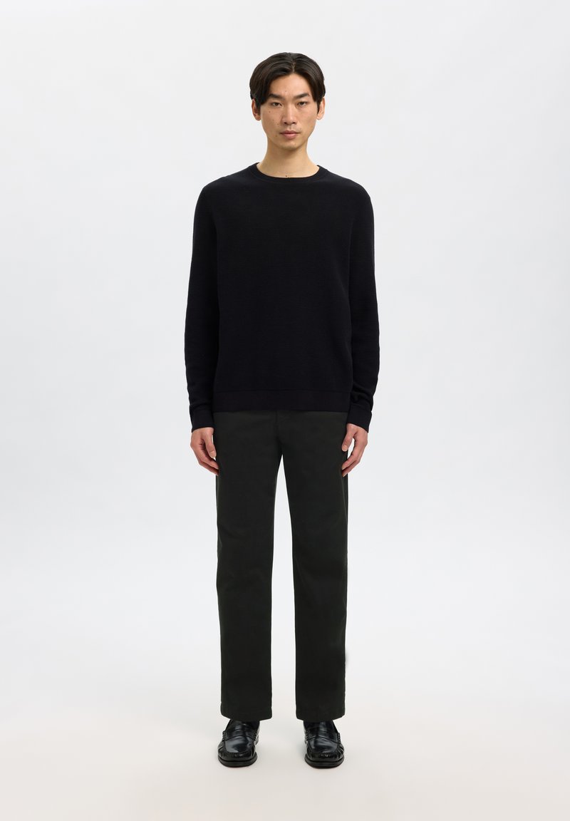 Ung mand står iført en sort langærmet sweater, mørke bukser og sorte sko mod en ensfarvet lys baggrund.