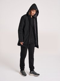 Giacca nera isolata con cappuccio, caratterizzata da una chiusura con zip e tasche laterali, abbinata a pantaloni neri e scarpe da ginnastica grigie.
