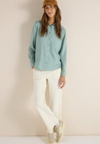 Femme portant une chemise boutonnée bleu clair, un pantalon ample crème, des baskets beiges et une casquette marron, debout contre un mur beige.