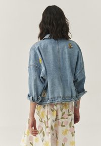 Leren denimjack in lichtblauw met opgerolde mouwen, geborduurde patches van gele bijen en een cropped ontwerp. Accenten zijn onder andere een label aan de zoom.
