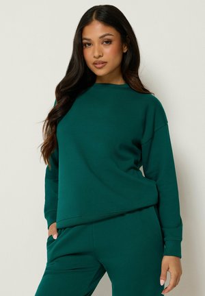 Kvinde med langt mørkt hår iført en mørkegrøn sweatshirt og matchende bukser, stående med den ene hånd i lommen mod en ensartet baggrund.
