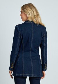 Blazer in denim blu scuro, con vestibilità sartoriale, bottoni a doppia fila sulle maniche e dettagli di cucitura visibili. Tessuto liscio.