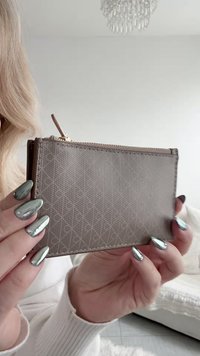 Γυναίκα με μεταλλικά ασημί μανικιούρ κρατάει πορτοφόλι με φερμουάρ σε απόχρωση ταουπ με υφή και διακριτικό ανάγλυφο σχέδιο σε εσωτερικό χώρο.