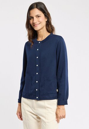 Femme aux cheveux châtain moyen portant un cardigan bleu marine boutonné avec des poches et un pantalon de couleur crème, souriant légèrement, sur un fond uni.