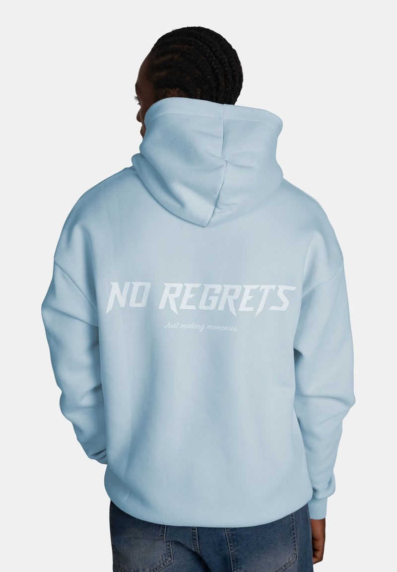 Personne aux cheveux tressés portant un sweat à capuche bleu clair avec les inscriptions "NO REGRETS" et "Just making memories" au dos.