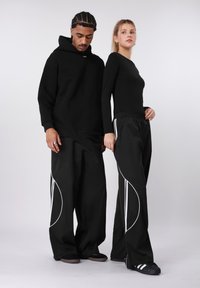 Fekete kapucnis pulóver elülső zsebbel és húzózsinórral, laza fekete nadrággal, fehér oldalsávokkal. Simább, puha anyagok.