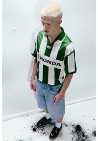 Groen en wit gestreepte polo met het "HONDA" logo, gecombineerd met lichtdenim shorts. Zwarte schoenen en zilveren sieraden accentueren de look.