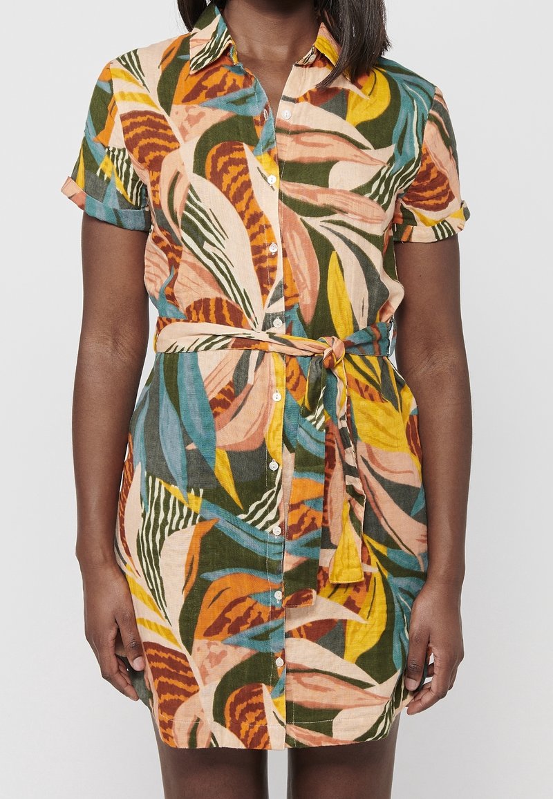Femme portant une robe boutonnée à manches courtes avec un motif abstrait de feuilles multicolores et une ceinture nouée à la taille.