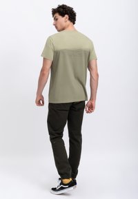 Olive green short-sleeve T-shirt with a gradient texture on the back, paired with dark green joggers and black sneakers with yellow socks.