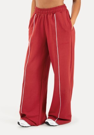 Rote weit geschnittene Hosen mit weißen vertikalen Streifen, getragen mit weißen Sneakern, Hände in den Taschen, elastischer Bund sichtbar.
