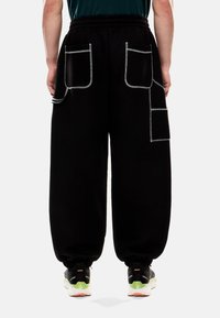 Pantaloni della tuta neri con una vestibilità ampia, caratterizzati da numerose tasche bordate da cuciture bianche e polsini elasticizzati alle caviglie.