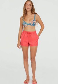 Naine kannab sinise lilleformaadi bikiinipluusi, korallivärvi nööriga lühikesi pükse ja vastavaid korallivärvi sussikesi, seistes ühe käega taskus.