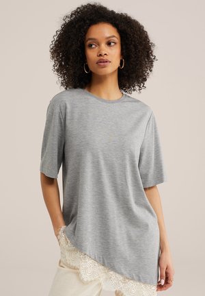 Kvinde med krøllet hår iført en løs grå T-shirt med asymmetrisk sømkant og blonder, beige bukser og guldringeøreringe.