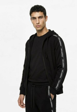 Jonge man draagt een zwarte zip-up hoodie en bijpassende broek met "KARL LAGERFELD" tekst langs de mouwen en broekspijpen.