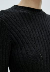 Zwarte geribbelde gebreide trui met een ronde hals, textuur en subtiel diagonaal stiksels patroon. Close-up van het mouwdetail.