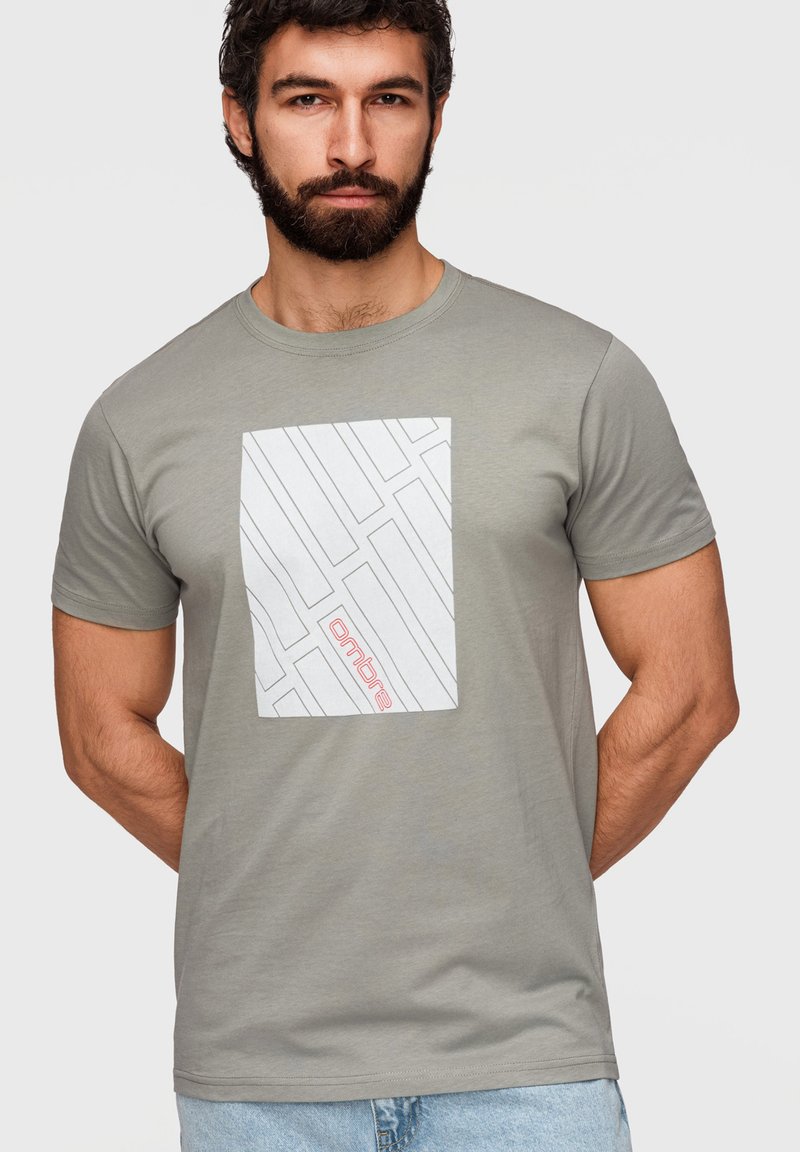 Homme barbu portant un t-shirt gris clair avec un motif géométrique blanc et un texte rouge « ombre », associé à un jean bleu clair.