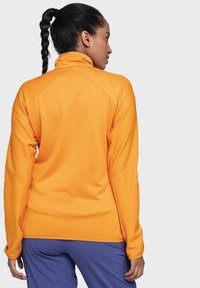 Orange Zip-Jacke mit gerippter Textur, entworfen mit hohem Kragen und langen Ärmeln, kombiniert mit lila Hose.