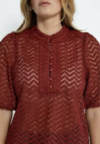 Bordo pitsikanga blouse, jossa on chevron-kuvio, korkea kaula-aukko ja nappilista. Lyhyet puffihihat ja läpinäkyvä rakenne.