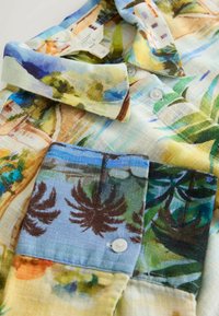 Cămașă din in cu mâneci scurte, având un imprimeu tropical colorat cu motive de palmieri, flori și peisaje. Nasturii sunt albi și rotunzi.