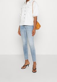 Camisa blanca de botones con mangas abullonadas, jeans ajustados de color azul claro y chanclas negras. Incluye un pequeño bolso naranja de hombro.