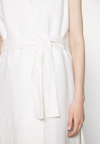 Ein weißes Leinen-Wickelkleid mit tiefem V-Ausschnitt, breitem Bindeband in der Taille und einem glatten, leicht strukturierten Stoff. Weicher Fall und minimalistisches Design.
