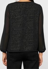 Zwarte blouse met een transparante achterkant en een textuurpatroon. Lange mouwen met een strakke manchette en een ronde halslijn.