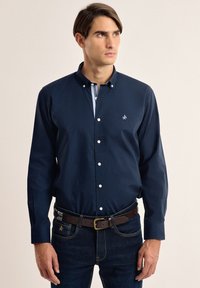 Camisa de botones azul marino con collar a rayas y logo en el pecho; combinada con jeans de mezclilla oscuros y un cinturón marrón. Textura suave.